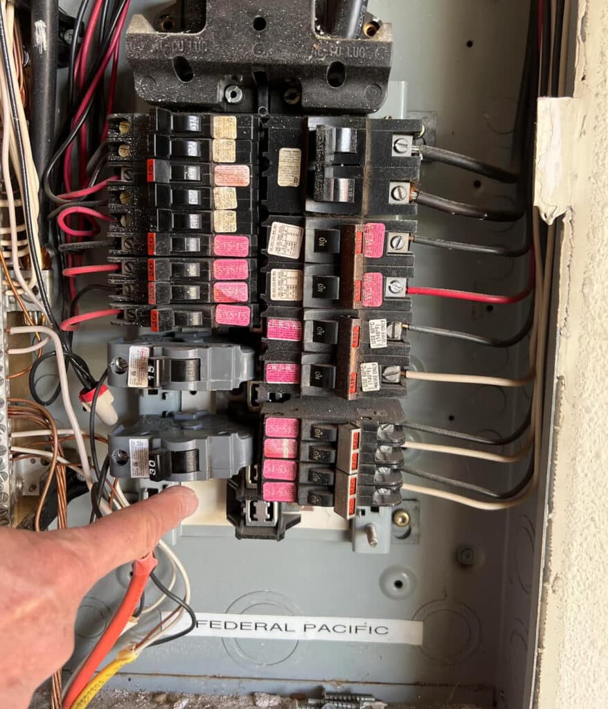 A hand points to breakers in an uncovered Federal Pacific panel with exposed wiring.